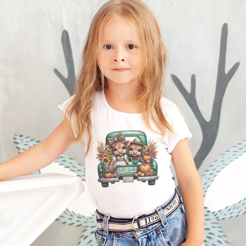 May include: A white t-shirt featuring a teal truck bed filled with pumpkins, sunflowers, and two cartoon cows. The text "fall fall" is printed on the truck. The girl is wearing blue jeans and a belt.