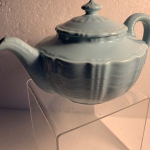 以下が含まれることがあります： フリル模様の蓋が付いた薄い青色のセラミック製のティーポット。ティーポットは透明なアクリル製の台座の上に置かれています。