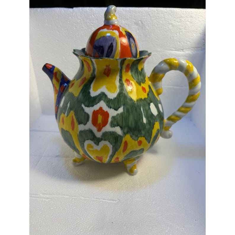 Pode incluir: Uma chaleira de cer&acirc;mica colorida com um design estampado em verde, amarelo, vermelho e branco. A chaleira tem uma al&ccedil;a torcida e tr&ecirc;s p&eacute;s. A tampa tamb&eacute;m &eacute; decorada com um padr&atilde;o colorido.