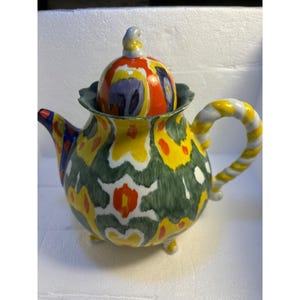Pode incluir: Uma chaleira de cer&acirc;mica colorida com um padr&atilde;o abstrato verde, amarelo, branco, vermelho e azul. A chaleira tem uma tampa com um bot&atilde;o e uma al&ccedil;a torcida.