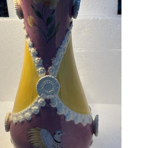 Puede incluir: Un jarr&oacute;n de cer&aacute;mica amarillo y morado con detalles blancos y un dise&ntilde;o de p&aacute;jaro. El jarr&oacute;n tiene una boca ancha y una base estrecha.