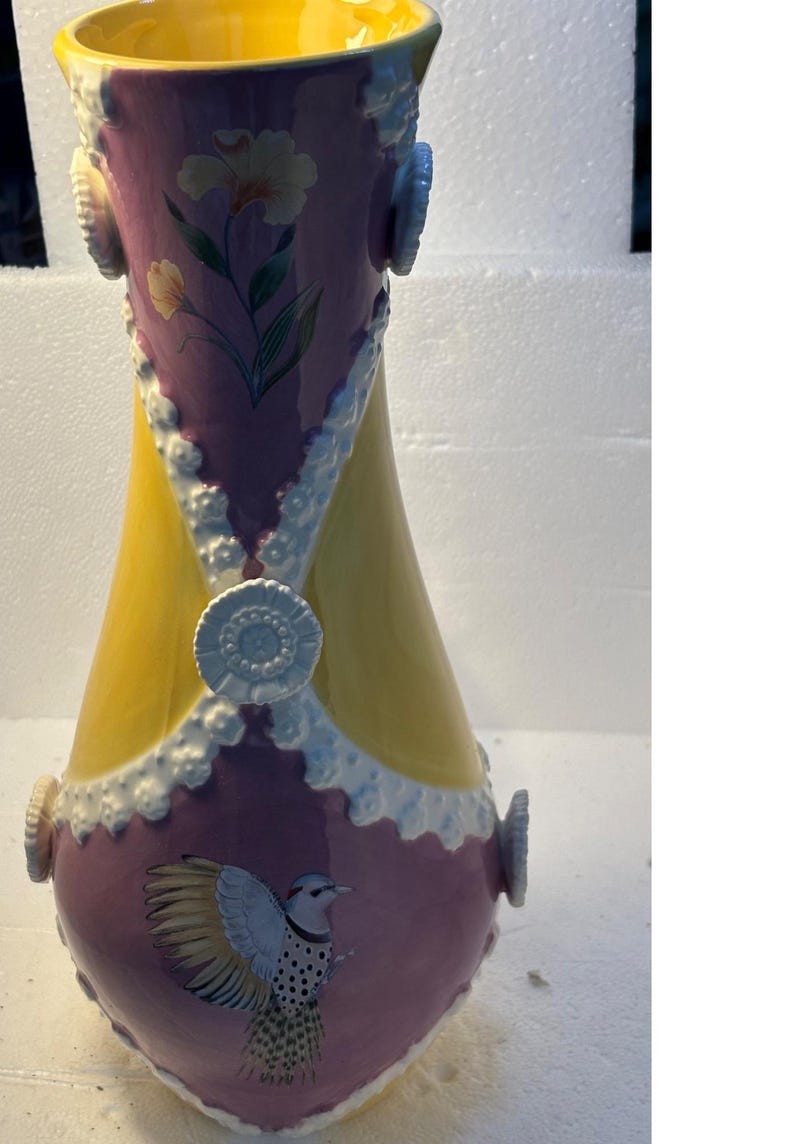 Puede incluir: Un jarr&oacute;n de cer&aacute;mica amarillo y morado con detalles blancos y un dise&ntilde;o de p&aacute;jaro. El jarr&oacute;n tiene una boca ancha y una base estrecha.
