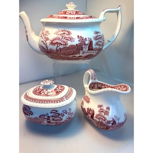 Puede incluir: Un juego de té de cerámica blanca con un patrón de paisaje rojo y blanco. El juego incluye una tetera, un azucarero con tapa y una jarra de crema. La tetera mide aproximadamente 25 cm de altura.