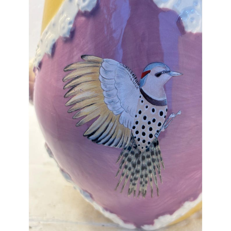 Puede incluir: Un jarr&oacute;n de cer&aacute;mica rosa con un dise&ntilde;o de p&aacute;jaro blanco y amarillo. El p&aacute;jaro tiene manchas negras y una cola larga.