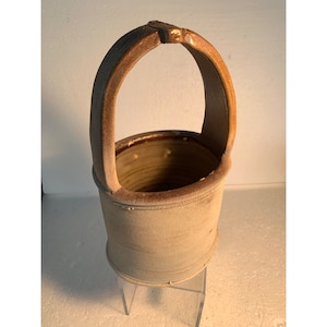 Puede incluir: Un macetero de cerámica con un asa marrón y un cuerpo gris claro. El macetero está sobre un soporte transparente.