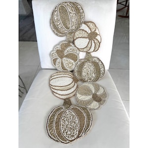 Può includere: Un runner da tavola decorativo con una disposizione verticale di sette applicazioni a forma di zucca. Ogni zucca è finemente lavorata con perline di vetro color oro metallizzato e bianco, creando una superficie testurizzata e brillante.