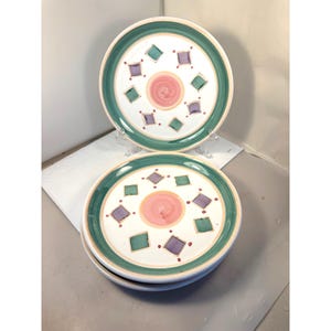 Può includere: Una pila di piatti rotondi in ceramica con un centro bianco caratterizzato da un motivo a spirale rosa, circondato da rombi alternati viola e verde acqua. Il bordo è decorato con un'ampia fascia verde e un sottile profilo color crema.