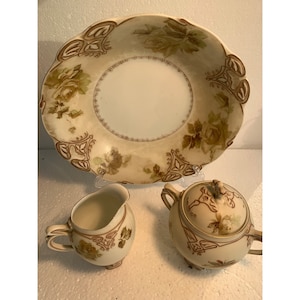 Pode incluir: Um conjunto de chá de porcelana vintage com um design floral. O conjunto inclui um açucareiro, um jarro de creme e uma tigela grande para servir. As peças são decoradas com rosas e folhas marrons e verdes sobre um fundo creme.
