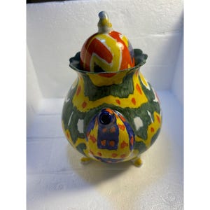 Pode incluir: Uma chaleira de cer&acirc;mica colorida com um padr&atilde;o geom&eacute;trico verde, amarelo, vermelho, azul e branco. A chaleira tem uma tampa com um padr&atilde;o semelhante e tr&ecirc;s p&eacute;s.