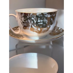 May include: White teacup and saucer set with gold trim. The cup and saucer feature a floral design in gold and silver. The teacup has a curved handle. The set is on a clear surface.