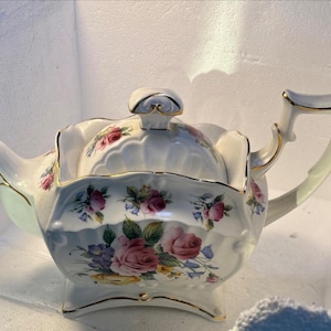 以下が含まれることがあります： 花柄と金色の縁取りが施された白いティーポット。ティーポットには装飾的なノブが付いた蓋と側面に注ぎ口があります。ティーポットはピンク、紫、緑の花で飾られています。