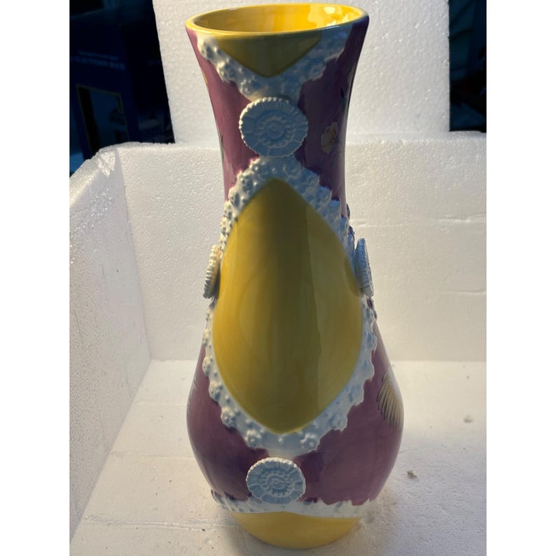 Puede incluir: Un jarr&oacute;n de cer&aacute;mica amarillo y morado con un dise&ntilde;o de encaje blanco y acentos circulares blancos. El jarr&oacute;n tiene una base ancha y un cuello estrecho.