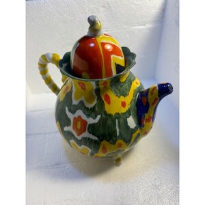Pode incluir: Uma chaleira de cer&acirc;mica colorida com um padr&atilde;o floral verde, amarelo, vermelho e branco. A chaleira tem uma tampa com um padr&atilde;o geom&eacute;trico em vermelho, laranja e azul. A chaleira tem tr&ecirc;s p&eacute;s e uma al&ccedil;a torcida.