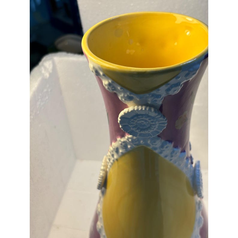 Puede incluir: Un jarr&oacute;n de cer&aacute;mica con un dise&ntilde;o amarillo, morado y blanco. El jarr&oacute;n tiene un borde festoneado y est&aacute; decorado con un ribete blanco de encaje y botones blancos.