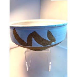 May include: A ceramic bowl with a light blue exterior and a white interior. The bowl features a brown, abstract design around the exterior. The bowl is sitting on a clear acrylic stand. The bowl is likely handmade.