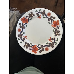 Puede incluir: Un plato de cerámica blanco con un diseño floral. El plato tiene un borde de flores naranjas y hojas verdes sobre un fondo azul oscuro.