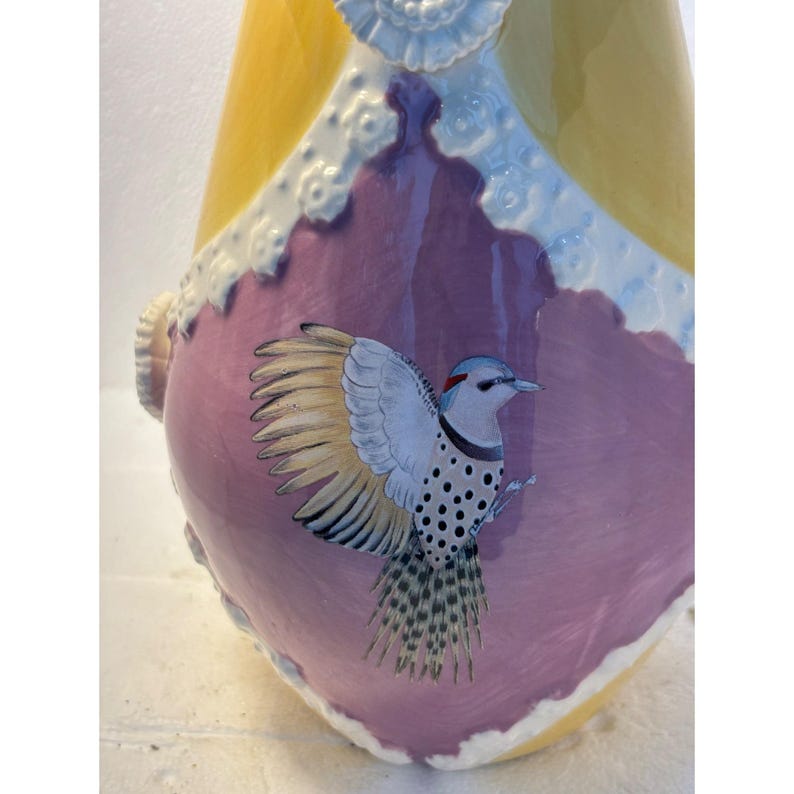 Puede incluir: Un jarr&oacute;n de cer&aacute;mica amarillo y morado con un dise&ntilde;o de p&aacute;jaro. El p&aacute;jaro est&aacute; pintado en tonos de azul, blanco y negro. El jarr&oacute;n tiene un dise&ntilde;o de encaje blanco alrededor de la parte superior.