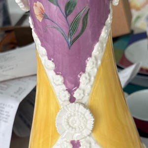 Puede incluir: Un jarr&oacute;n de cer&aacute;mica con un dise&ntilde;o amarillo y morado. El jarr&oacute;n tiene un ribete de encaje blanco y presenta un p&aacute;jaro y flores pintadas.