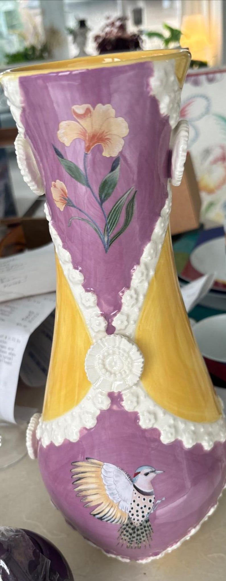 Puede incluir: Un jarr&oacute;n de cer&aacute;mica con un dise&ntilde;o amarillo y morado. El jarr&oacute;n tiene un ribete de encaje blanco y presenta un p&aacute;jaro y flores pintadas.