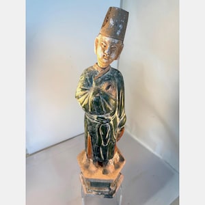 May include: A standing ceramic figurine of a person wearing a tall, weathered tan hat and a dark green glazed robe with orange accents. The figure has a light-colored face and stands on a hexagonal base, showing signs of age and surface wear.