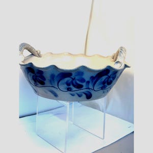 May include: A beige ceramic bowl with a scalloped rim and two side handles. The exterior features a hand-painted blue floral pattern with swirling vines. The bowl has a textured, speckled finish and is elevated on a clear acrylic display stand.
