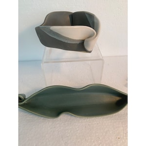 Puede incluir: Dos cuencos de cerámica, uno es de color gris oscuro con detalles blancos y el otro es de color verde claro. Ambos cuencos tienen una forma única y orgánica.