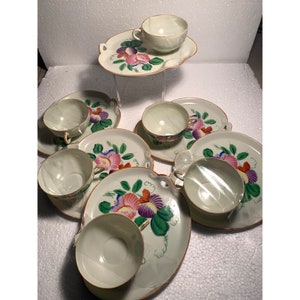 May include: A set of six vintage teacups and saucers with floral designs. The cups and saucers are white with a green, pink, and purple floral pattern. The saucers have a scalloped edge.