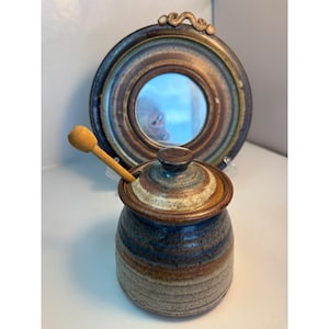 Könnte beinhalten: Handgemachter Keramik-Honigtopf mit einem hölzernen Honiglöffel. Der Topf ist zylindrisch mit Deckel und in Blau-, Braun- und Beigetönen glasiert. Im Hintergrund ein dekorativer runder Spiegel mit braunem Rahmen.