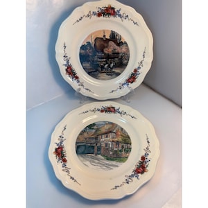 Puede incluir: Dos platos decorativos de color crema con bordes festoneados. Cada plato presenta una imagen central de una escena de pueblo europeo. Los bordes están adornados con patrones florales y de vid en rojo, azul y verde.