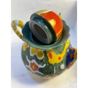 Pode incluir: Uma chaleira verde e amarela com um padr&atilde;o floral e uma tampa colorida. A tampa est&aacute; aberta e o interior da chaleira &eacute; vis&iacute;vel.