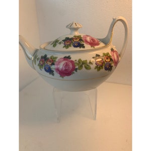 Puede incluir: Tetera blanca con un diseño floral con rosas rosas, rojas y azules y otras flores. La tetera tiene un borde dorado alrededor del borde y una perilla dorada en la tapa.