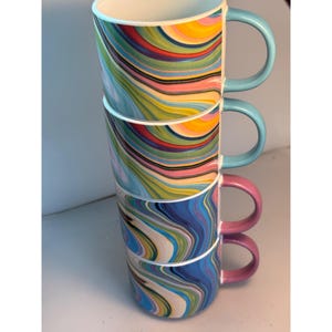 Puede incluir: Cuatro tazas de cerámica apiladas con patrones coloridos y arremolinados en tonos de azul, amarillo, rojo y verde. Cada taza tiene un asa de color sólido: azul claro, rosa y azul claro. Las tazas son blancas por dentro.