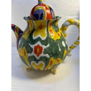 Pode incluir: Uma chaleira de cer&acirc;mica colorida com um padr&atilde;o floral verde, amarelo, vermelho e branco. A chaleira tem uma tampa com um padr&atilde;o semelhante e uma al&ccedil;a torcida.