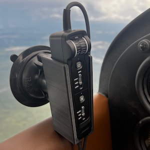 May include: A black aviation headset with a microphone and a control panel with buttons for volume, noise cancellation, and other settings. The headset is mounted on a suction cup mount on a dashboard.