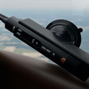 May include: A black, handheld audio recorder with a suction cup mount. The recorder has several buttons and a small screen. The recorder is attached to a window in an airplane.