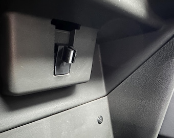 May include: A close-up of a black plastic latch or release mechanism, likely for a car door or compartment. The latch is in a recessed area and appears to be in the closed position.