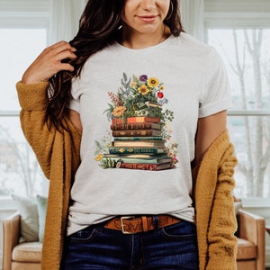 May include: A white t-shirt with a graphic of a stack of books with flowers and greenery. The books are vintage and have different colored spines. The flowers are in shades of pink, yellow, and white.