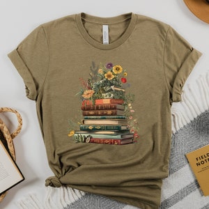 May include: A green t-shirt with a graphic of a stack of books with flowers and greenery on top. The books have colorful spines and are stacked in a pile.