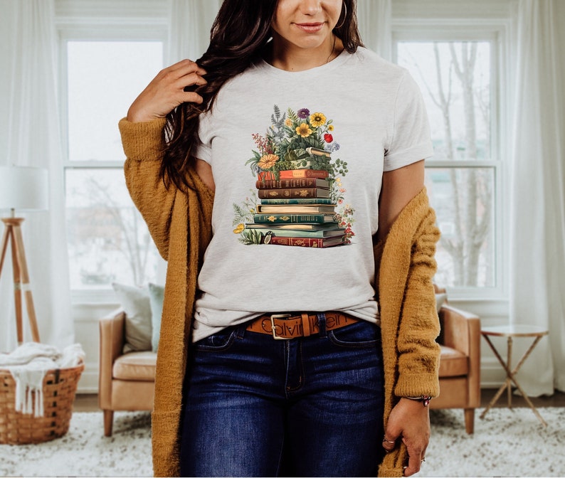 May include: A white t-shirt with a graphic of a stack of books with flowers and greenery. The books are vintage and have different colored spines. The flowers are in shades of pink, yellow, and white.