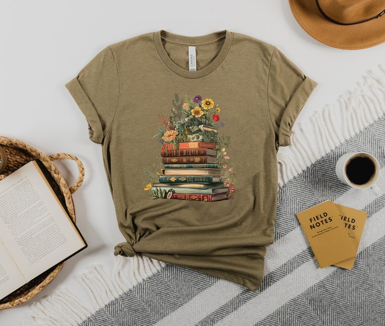 May include: A green t-shirt with a graphic of a stack of books with flowers and greenery on top. The books have colorful spines and are stacked in a pile.