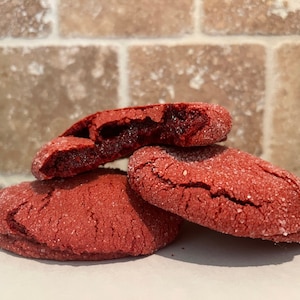 May include: Three red velvet cookies, one with a bite taken out, on a white surface. The cookies are covered in sugar crystals.