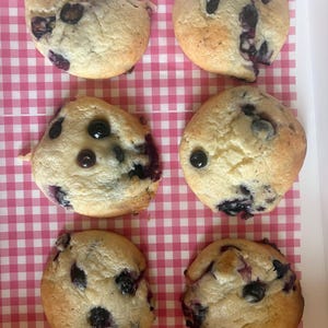 May include: Six round blueberry muffins arranged in two columns. Each muffin has a light golden-brown baked exterior with visible dark blue berries embedded throughout the surface. The muffins are placed on a white surface with a pink gingham pattern.