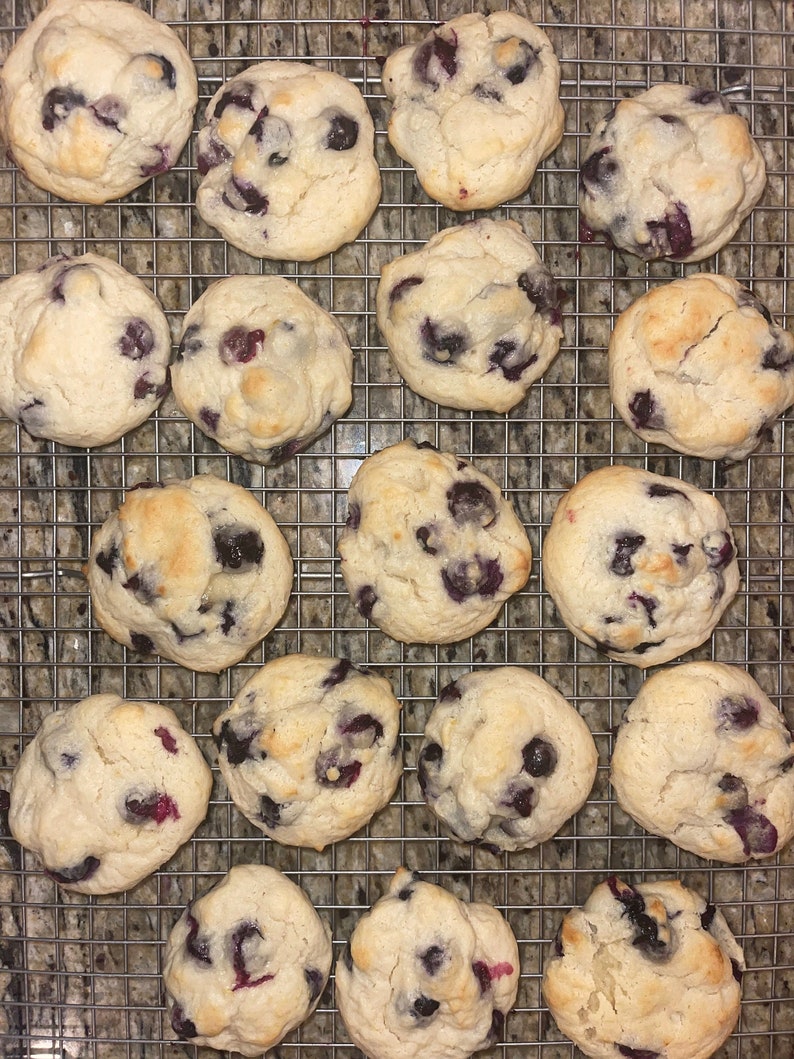 May include: A cooling rack with 18 blueberry cookies. The cookies are light brown and have a slightly golden hue. The blueberries are scattered throughout the cookies.