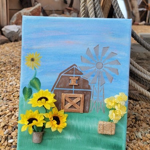 K&ouml;nnte beinhalten: Ein blaues Leinwandbild mit einem braunen Scheunen, einer Windm&uuml;hle und gelben Sonnenblumen. Die Scheune hat eine Holzt&uuml;r und ein Holzfenster. Die Sonnenblumen stehen in einem kleinen gr&uuml;nen Topf. Das Bild ist auf einem Hintergrund aus braunen Steinen.