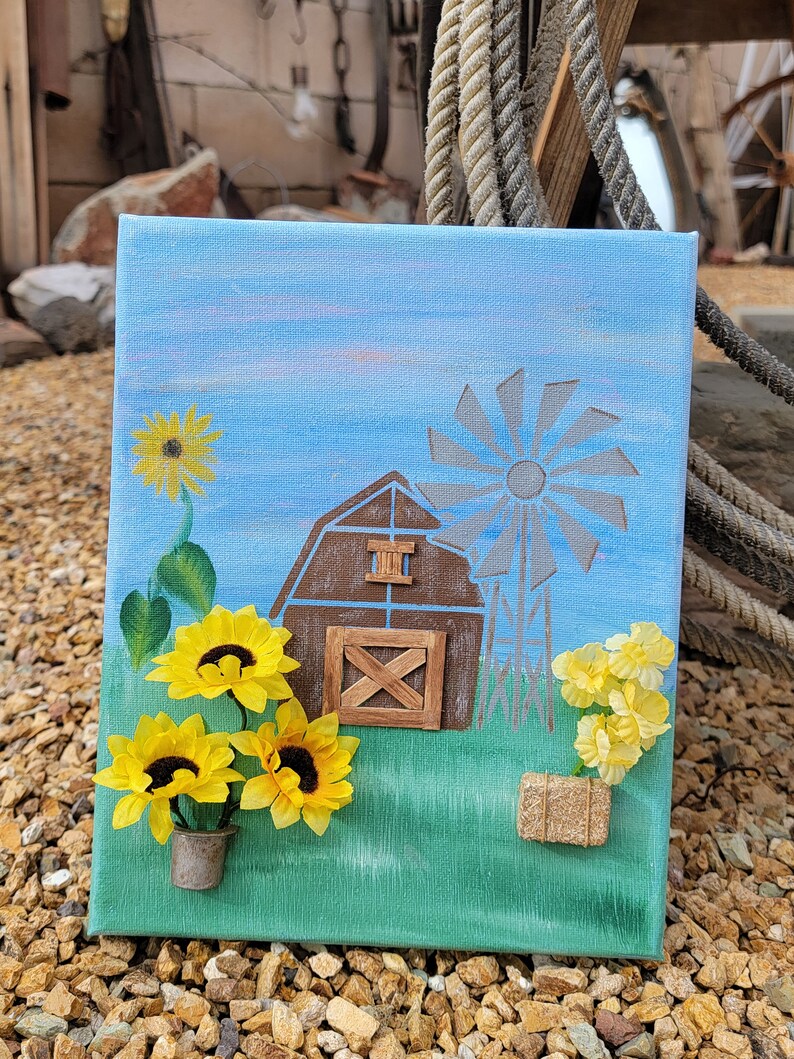 K&ouml;nnte beinhalten: Ein blaues Leinwandbild mit einem braunen Scheunen, einer Windm&uuml;hle und gelben Sonnenblumen. Die Scheune hat eine Holzt&uuml;r und ein Holzfenster. Die Sonnenblumen stehen in einem kleinen gr&uuml;nen Topf. Das Bild ist auf einem Hintergrund aus braunen Steinen.