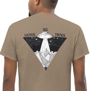 Peut inclure: Un t-shirt beige avec un motif noir et blanc d'un OVNI volant au-dessus de montagnes. Le motif comprend également le texte "Leave No Trace" dans une forme triangulaire.