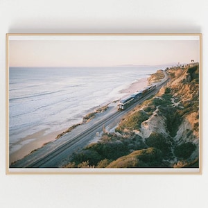 May include: A train travels along a coastal track with the ocean in the background. The train is on a cliff overlooking the beach. The sky is a light blue with a hint of pink.