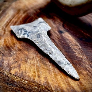 May include: A gray, hand-crafted arrowhead with a textured surface. The arrowhead has a triangular shape with a sharp point and a wider base. The stone artifact is displayed on a wooden surface.