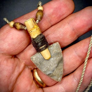 May include: A gray arrowhead pendant on a brown cord necklace with wooden beads. The arrowhead is attached to a small wooden cylinder wrapped with brown string.