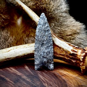 May include: A gray, hand-knapped arrowhead with a textured surface. The arrowhead is displayed with a deer antler and fur background. The arrowhead is approximately 3 inches long.